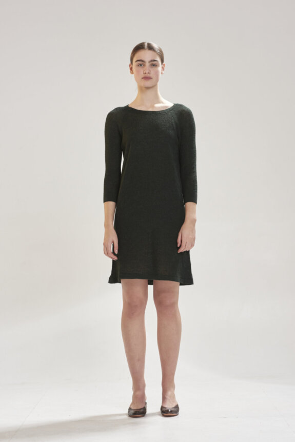 A woman wearing a dark green dress stands against a plain background.