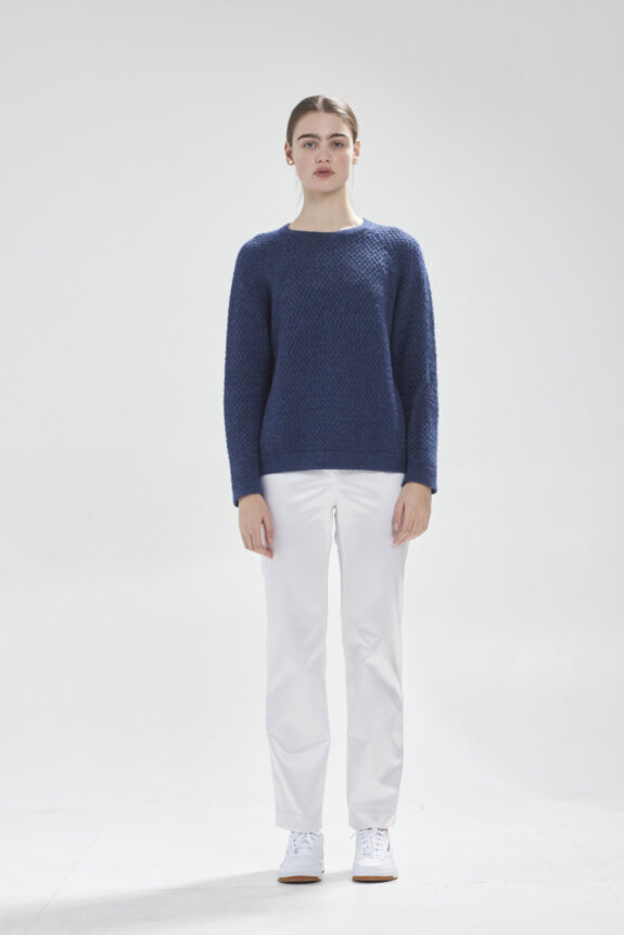 A person wearing a blue sweater, white pants, and white sneakers standing against a plain background.