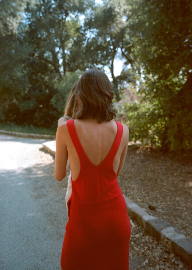 A woman in a red dress with an open back walks down a sunlit path surrounded by trees.