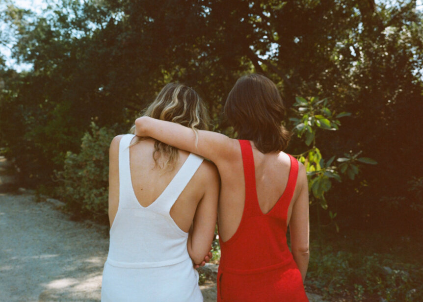 Two people with their arms around each other, one in a white dress and the other in a red dress, walking in a garden.