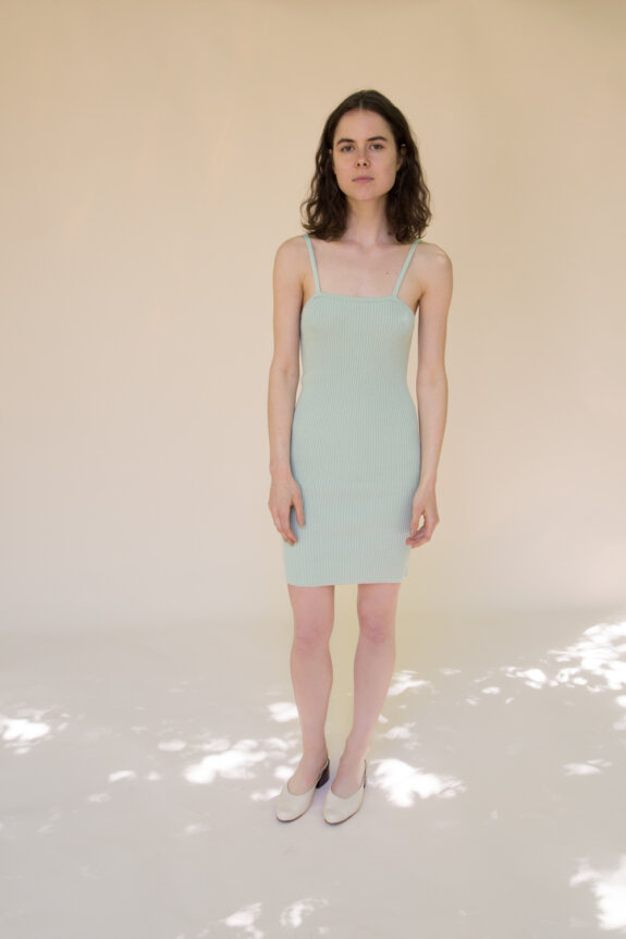 A woman is standing in a light green fitted dress and white shoes against a beige background.