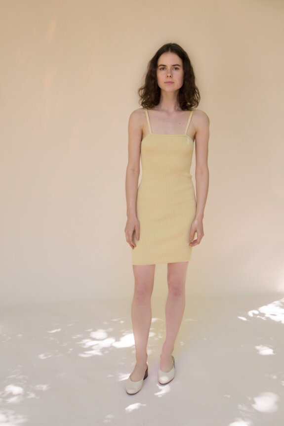A woman in a yellow ribbed dress and white shoes stands against a light beige background.