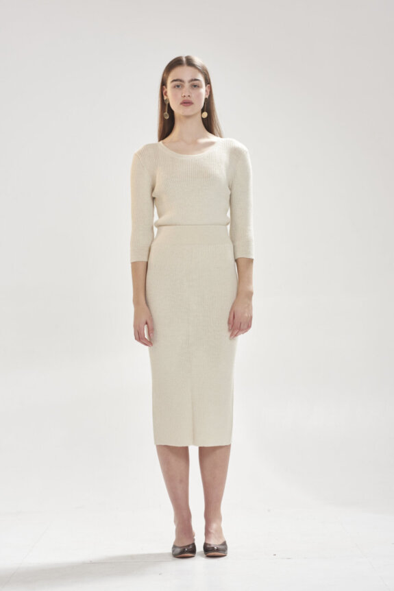 A woman in a beige knit dress stands against a plain background.