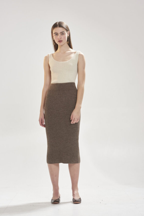 A woman is wearing a cream tank top and a brown knit skirt, standing against a plain background.