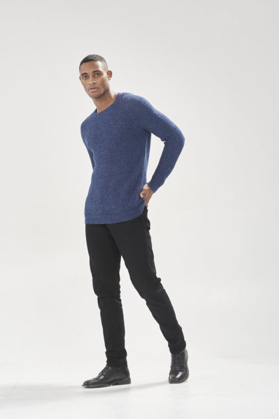 A man wearing a blue sweater and black pants poses in a studio setting.