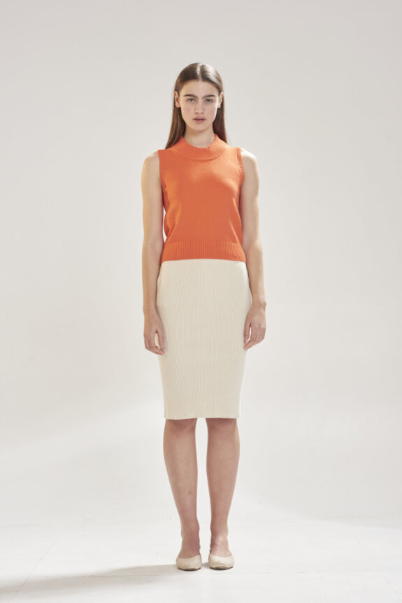 A woman is wearing an orange sleeveless top and cream pencil skirt with beige flats, standing against a plain background.
