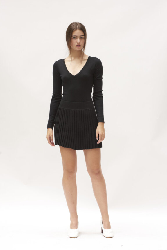 A woman is wearing a black V-neck long-sleeve top and pleated skirt with white shoes.