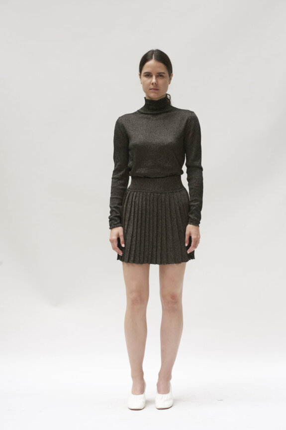 A woman is wearing a black turtleneck and pleated skirt with white shoes against a plain background.