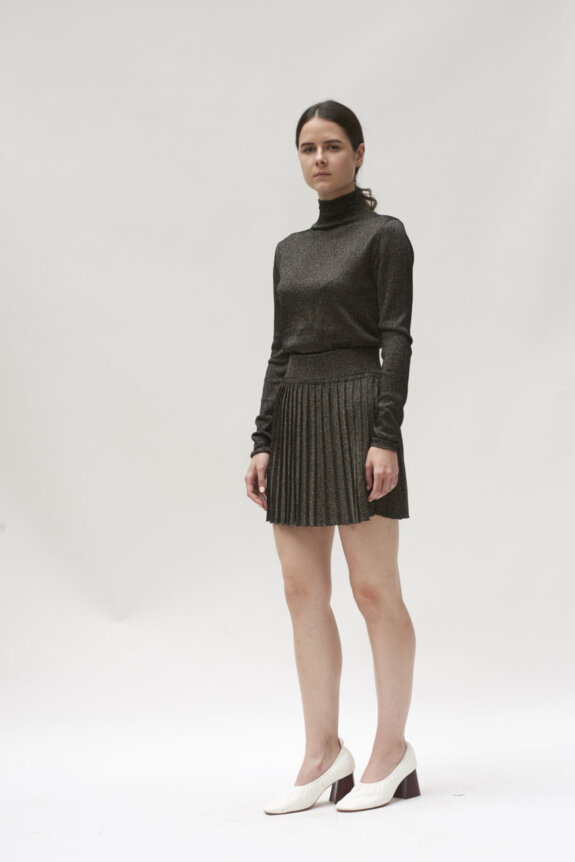 A woman wearing a dark turtleneck top, pleated skirt, and white heels stands against a plain background.