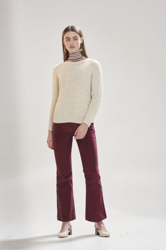 A woman wearing a cream sweater, striped turtleneck, red pants, and beige heels standing in a studio.