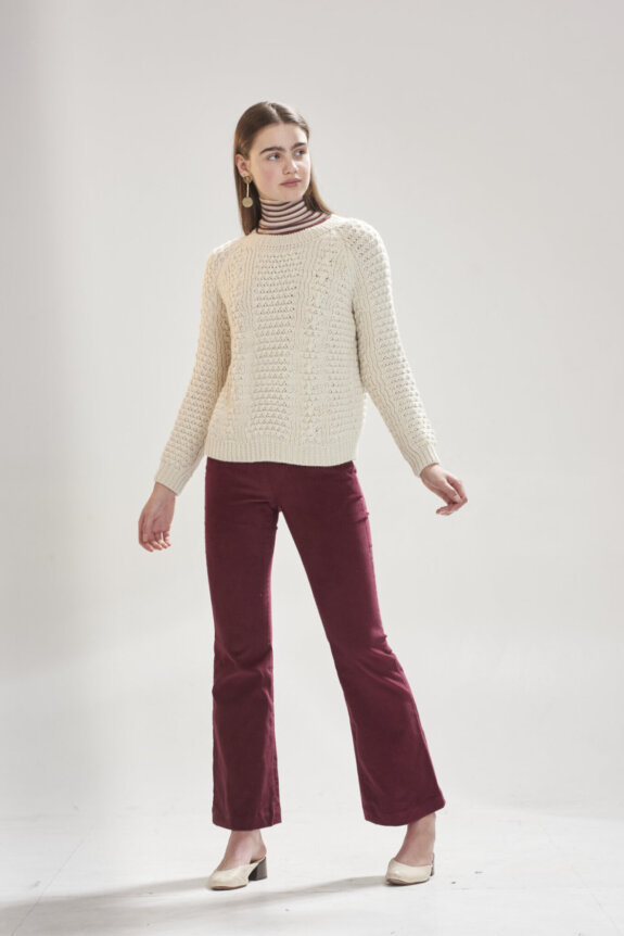 A woman wearing a cream knitted sweater, striped turtleneck, red pants, and white shoes poses against a plain background.