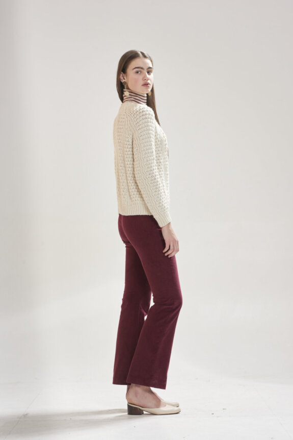 A woman in a cream sweater, striped turtleneck, and burgundy pants posing sideways.