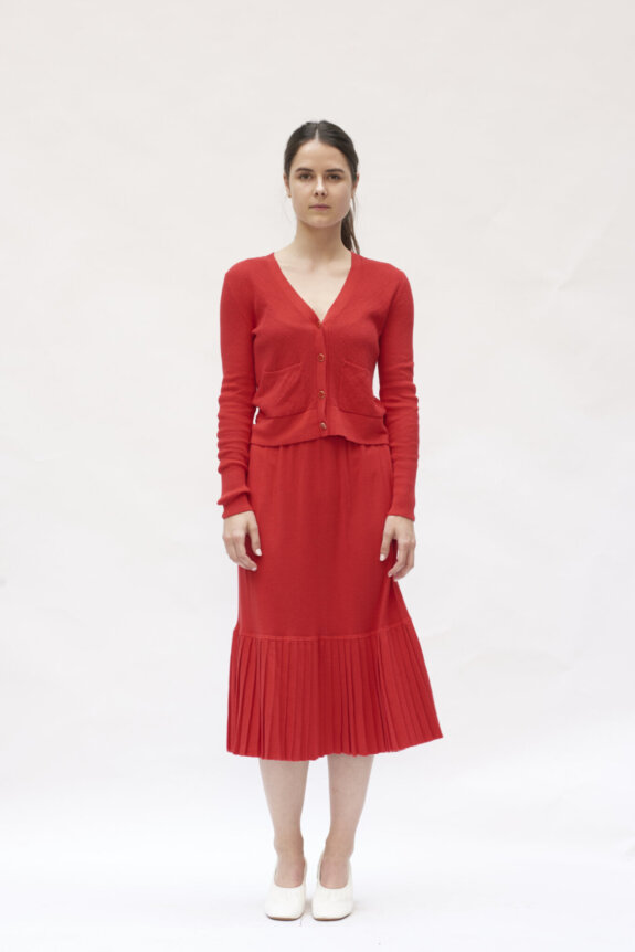 A woman wearing a red dress and cardigan stands against a plain background.