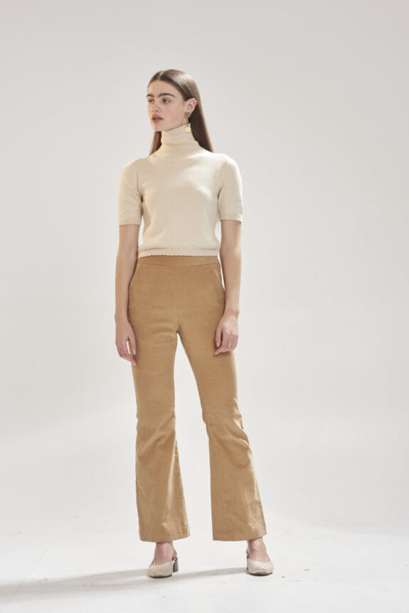 A woman wearing a beige turtleneck and tan pants stands in a neutral, well-lit space.