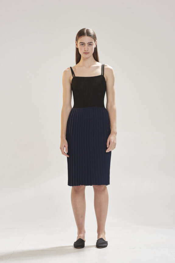 A woman in a black and navy ribbed dress with double straps stands against a plain background.