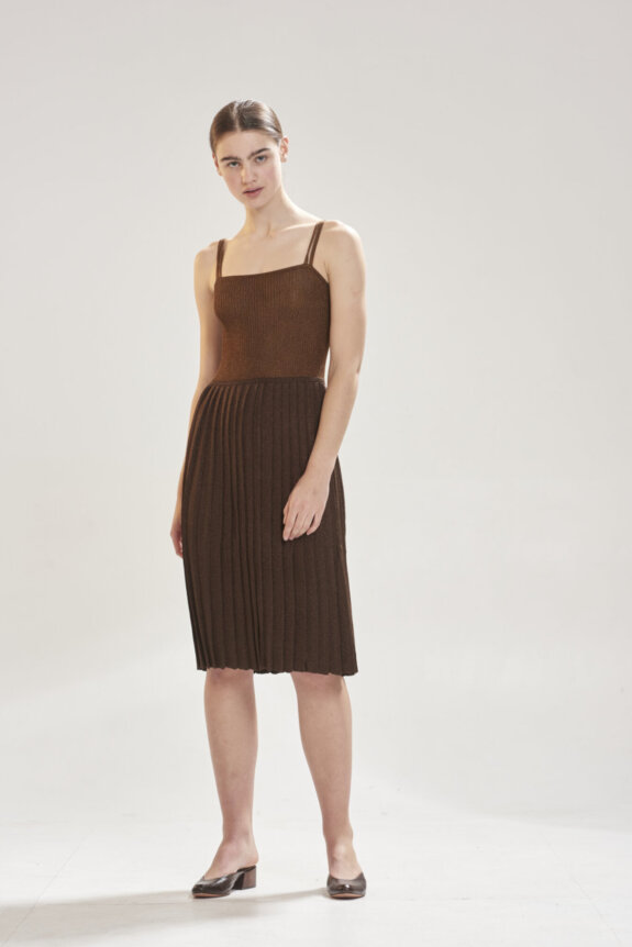 A woman wearing a brown pleated dress and brown shoes stands in a neutral setting.