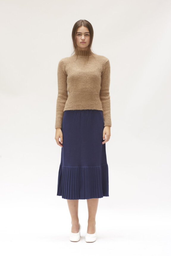A woman wearing a beige sweater, navy pleated skirt, and white shoes, standing against a plain background.