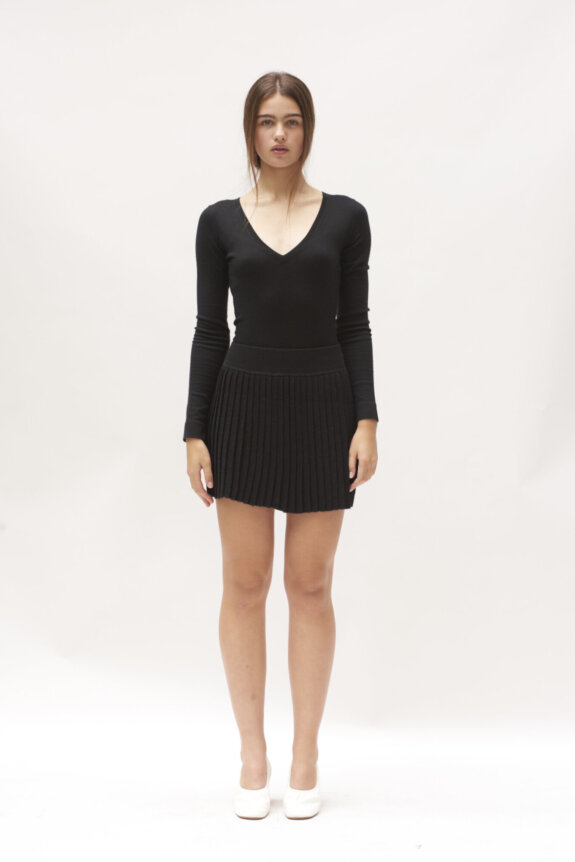 A woman stands wearing a black V-neck top and pleated skirt with white shoes.