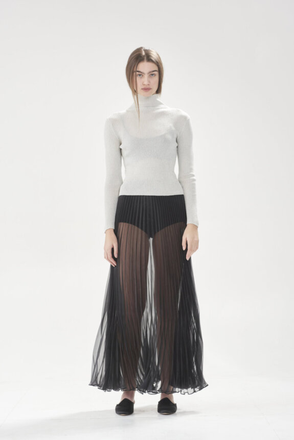A model wearing a white turtleneck and a sheer black pleated skirt stands in a neutral setting.