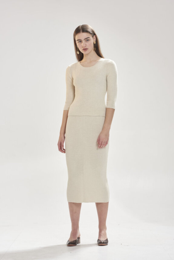 A woman is wearing a cream-colored knit top and skirt ensemble with hoop earrings and dark heels.