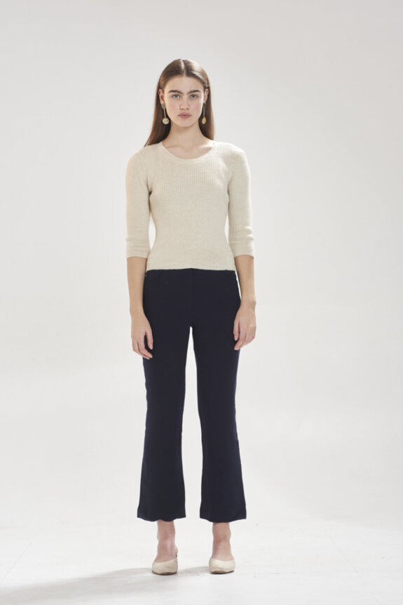 A woman in a cream sweater and black pants standing against a plain background.