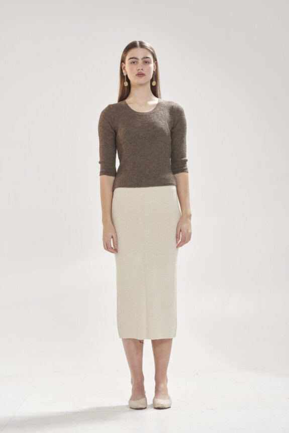 A woman wearing a brown top and cream skirt stands against a plain background.