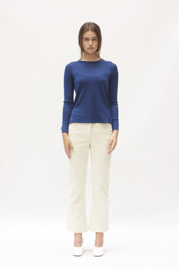 A woman standing in a blue long-sleeve shirt and cream pants against a white background.