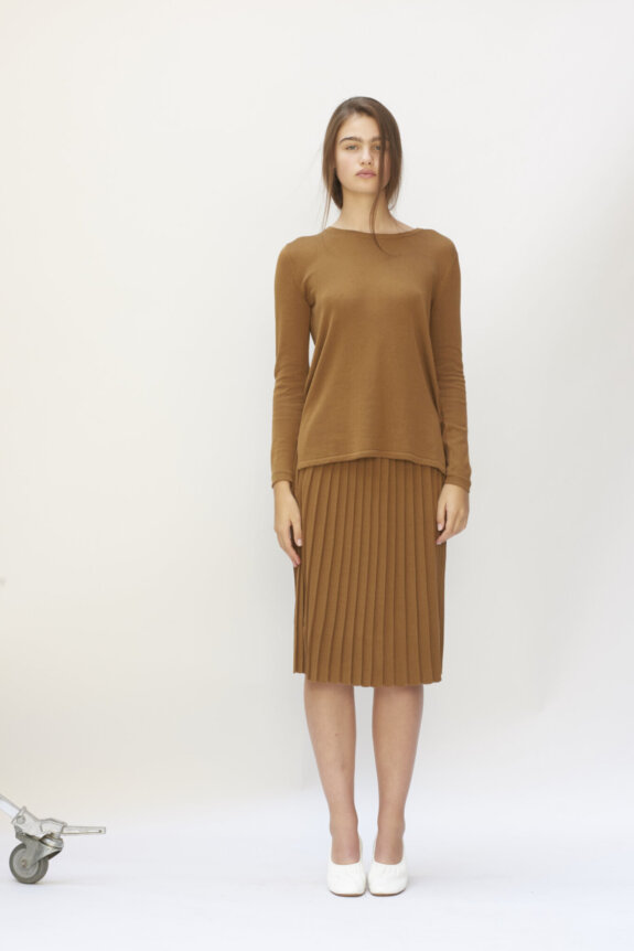 A person wearing a brown long-sleeve top and pleated skirt with white shoes against a plain background.