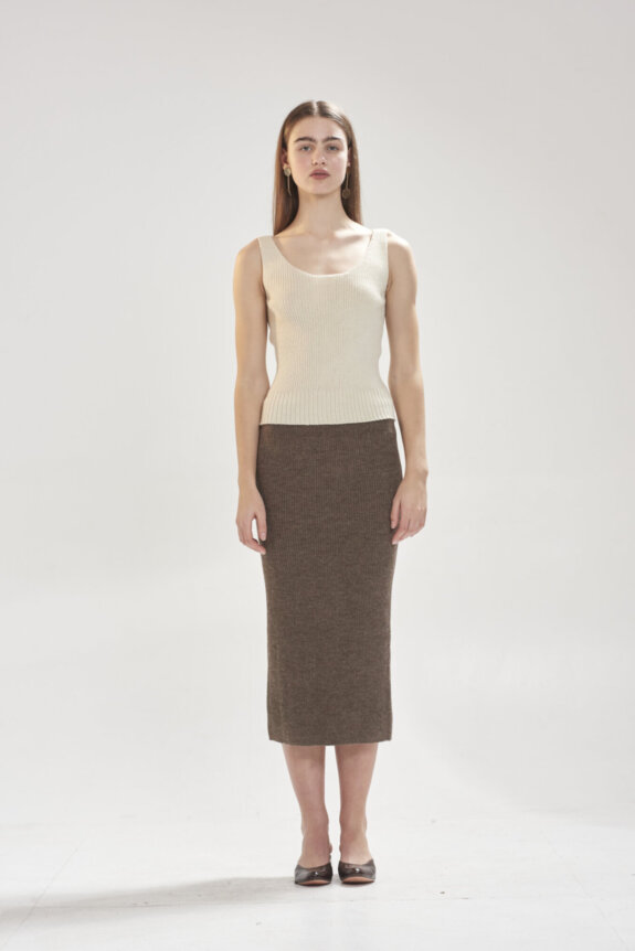 A woman standing in a fitted sleeveless top and a long brown skirt.