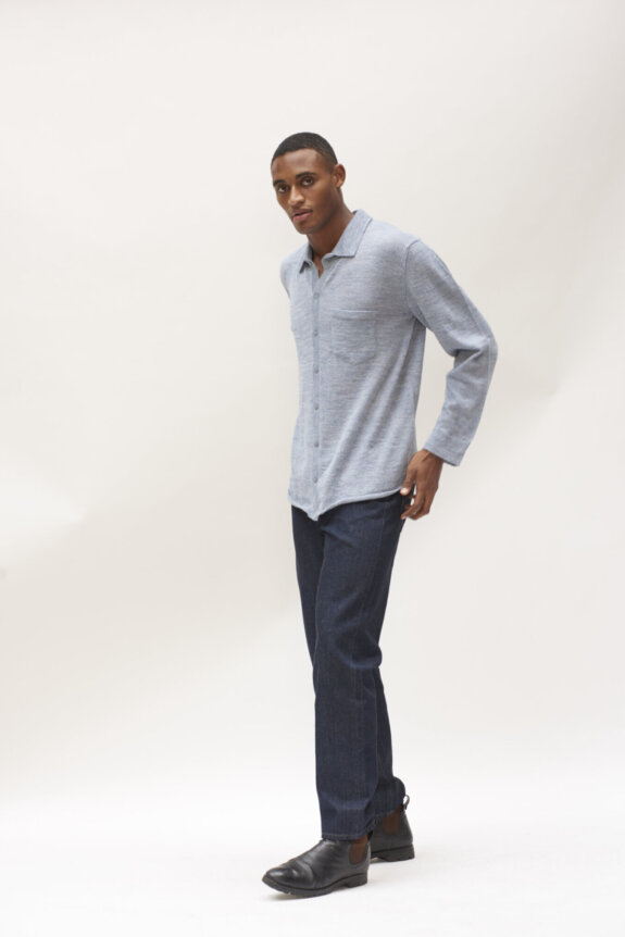 A man wearing a gray shirt, dark jeans, and black shoes poses in front of a plain background.