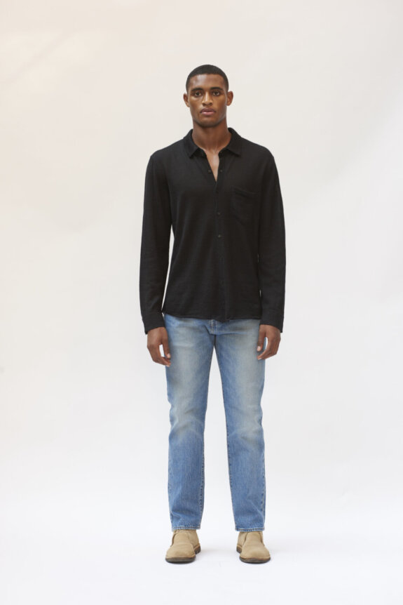 A man wearing a black shirt, blue jeans, and beige shoes standing against a plain background.