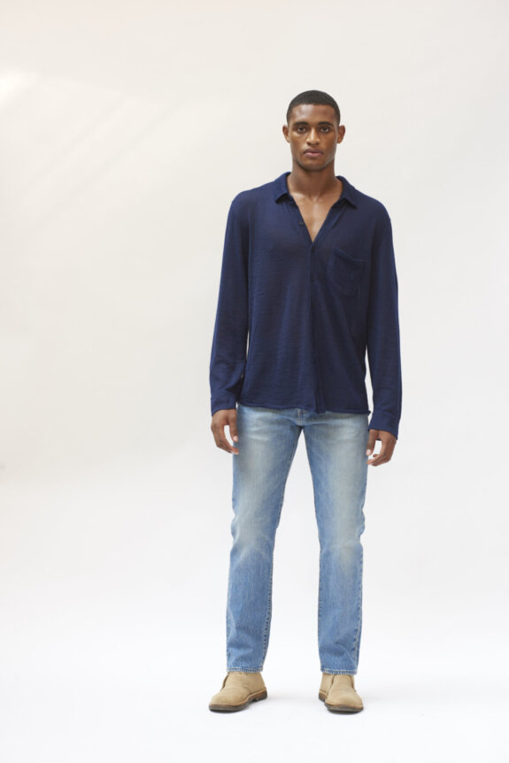 A man wearing a navy shirt and blue jeans stands against a plain white background.