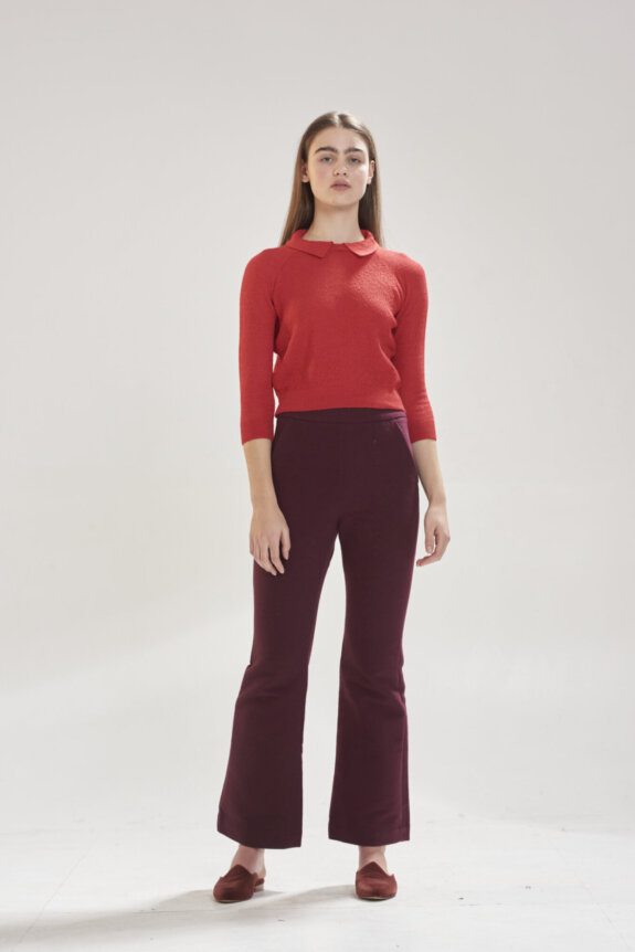 A person is standing wearing a red sweater and dark purple pants in a studio setting.