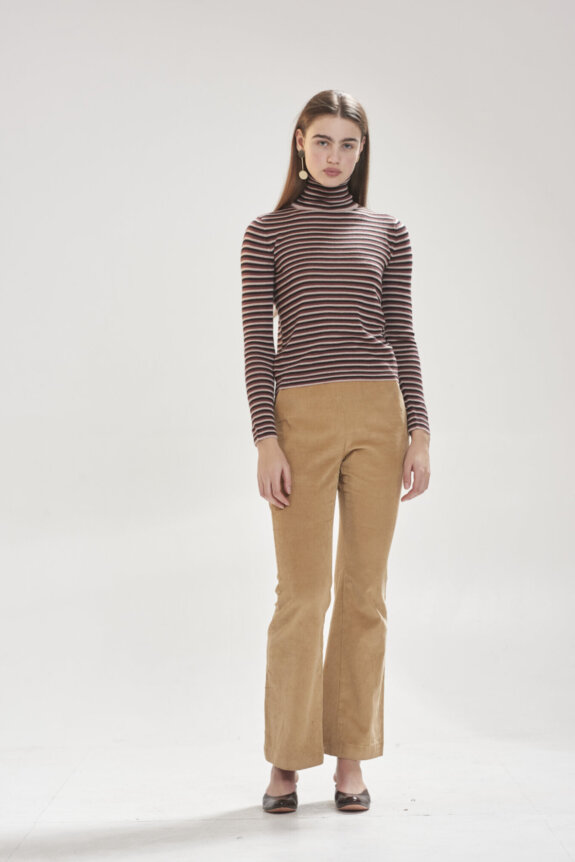 A person wearing a striped turtleneck and tan pants standing against a plain background.