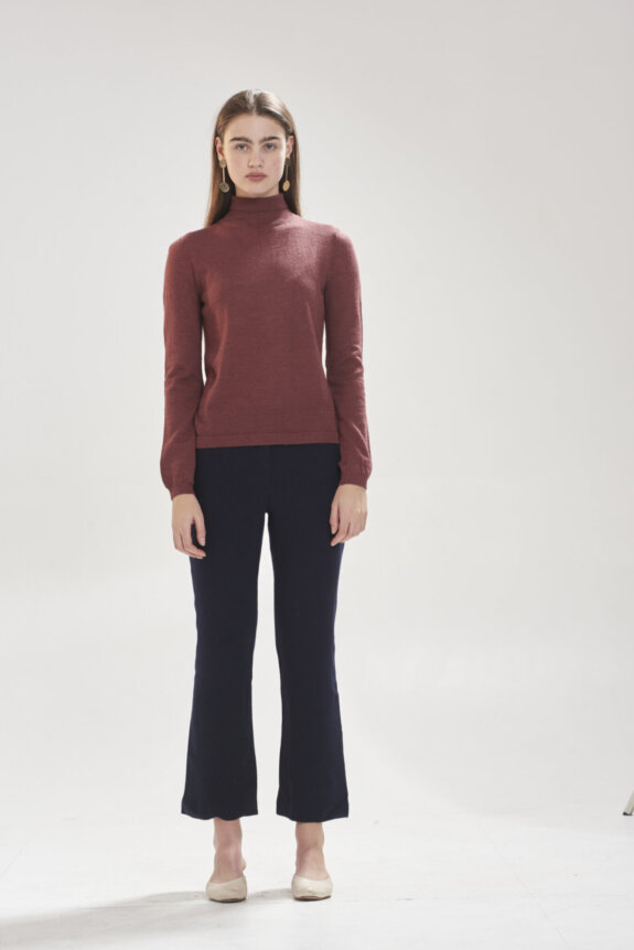 A woman in a red sweater and black pants stands against a neutral background.