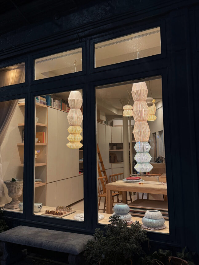 A warmly lit interior viewed through a window, showcasing stacked, textured pendant lights and neatly arranged shelves.