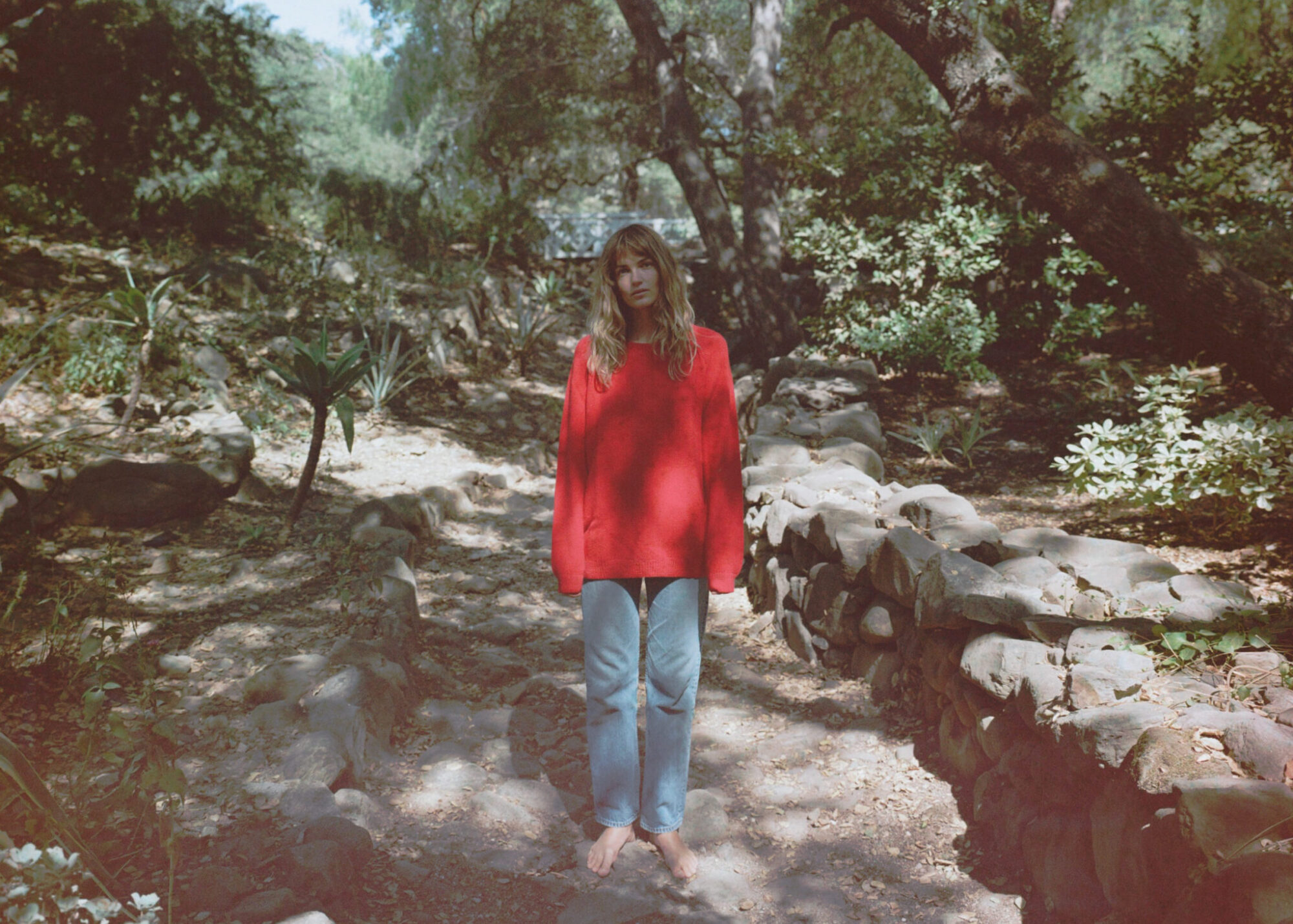 A person in a red sweater and jeans stands barefoot on a stone path in a wooded area.