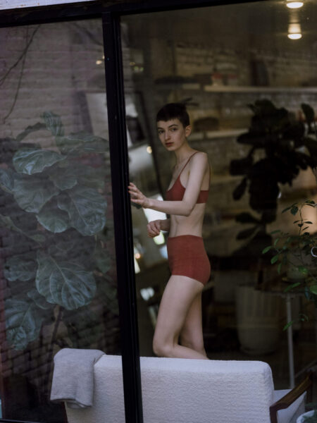 A person in red underwear stands by a window in a dimly lit room with plants.