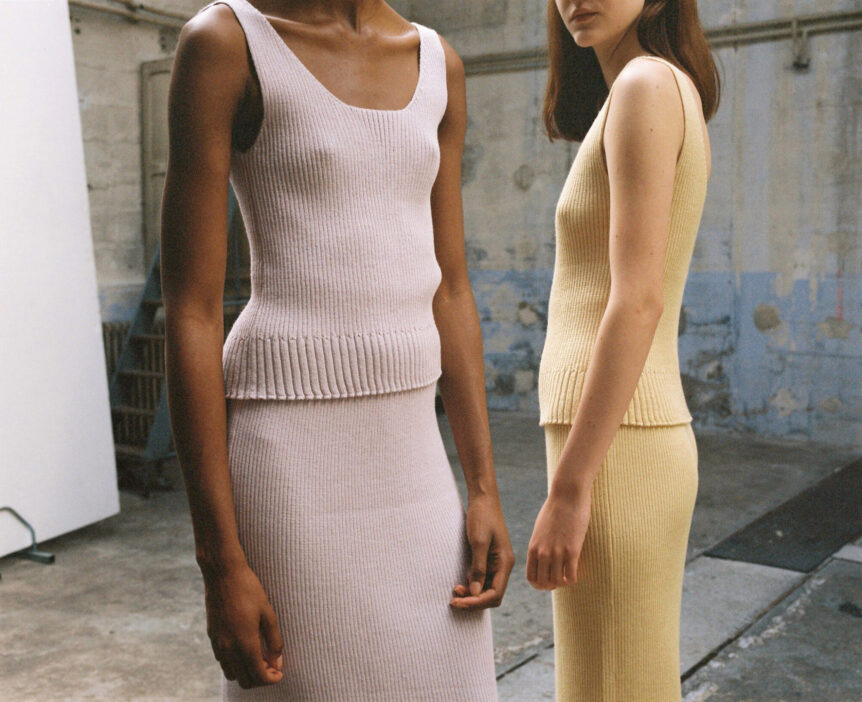 Two models wearing ribbed knit tank tops and skirts in a textured industrial setting.