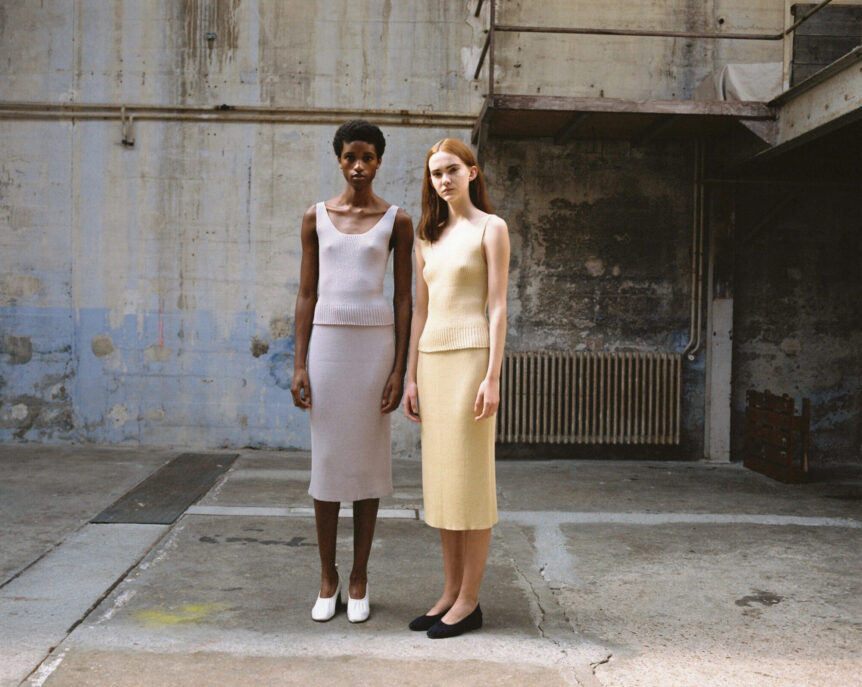 Two models wearing minimalist knit outfits stand in an industrial setting.