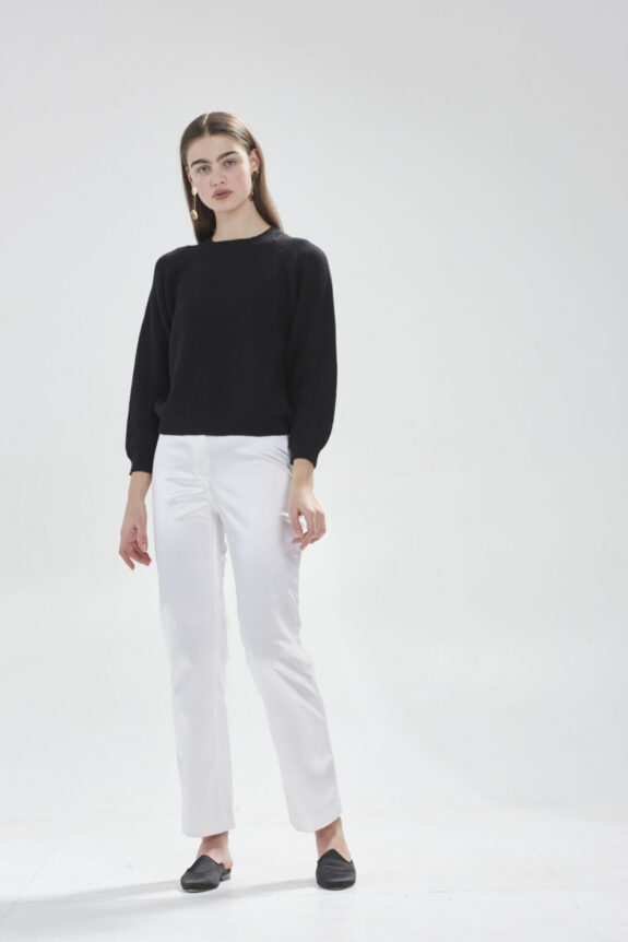 A woman wearing a black sweater, white pants, and black shoes, standing against a plain white background.