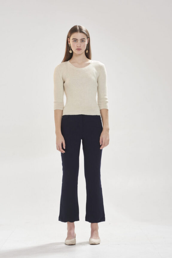 A woman in a light sweater and dark pants standing in a minimalist setting.