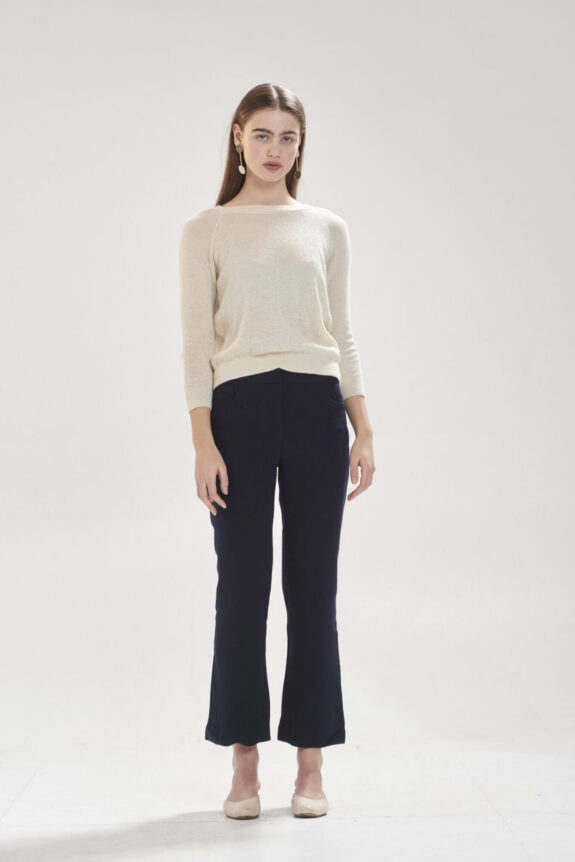 A woman wearing a light sweater and dark pants stands against a plain background.