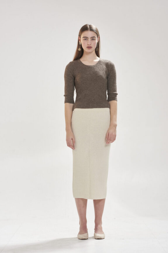 A person wearing a brown sweater and cream skirt standing against a plain white background.