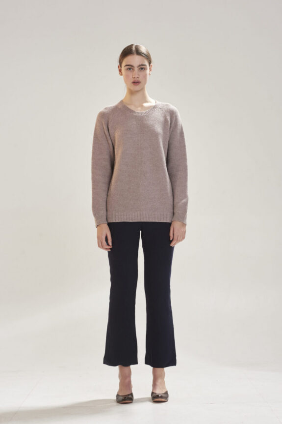 A person wearing a light brown sweater and dark pants standing in a neutral setting.