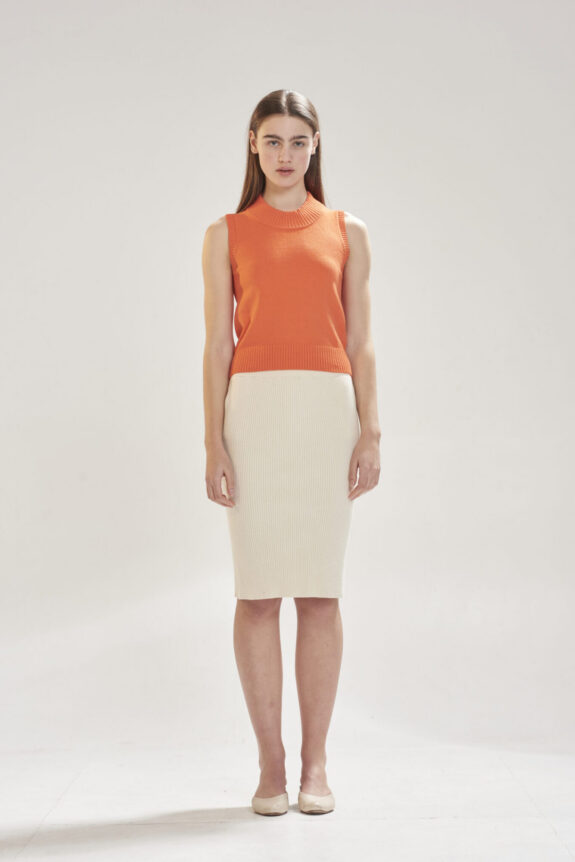 A woman wearing an orange sleeveless top and cream ribbed skirt stands against a plain background.