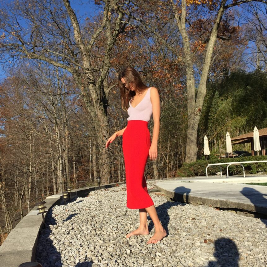 A woman stands barefoot on a rocky path wearing a pink top and red skirt.
