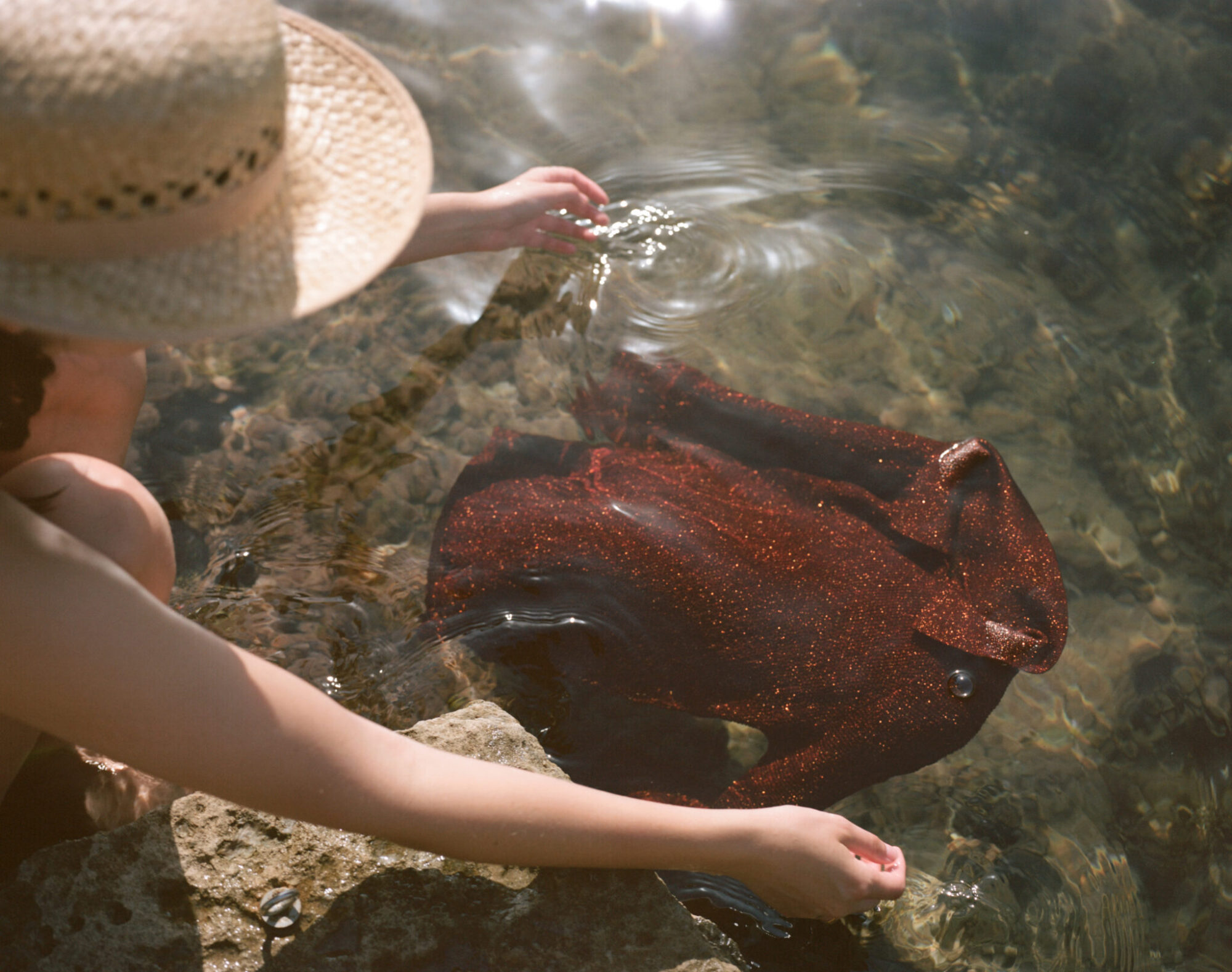 A person in a straw hat washes a glittery red garment in a clear body of water.