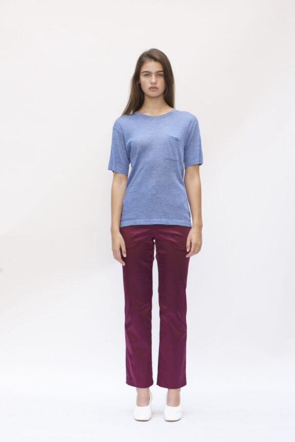 A woman wearing a blue t-shirt and burgundy pants standing against a plain background.