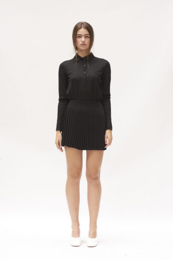 A woman wearing a black collared shirt and pleated skirt with white shoes.